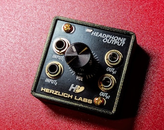 May include: Black headphone amplifier with gold-coloured input and output jacks. The amplifier has a knob labelled "VOL" and the text "OHP HEADPHONE OUTPUT" and "HERZLICH LABS".