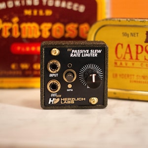 May include: A black and gold Herzlich Labs OHP Passive Slew Rate Limiter. The device has input and output jacks, a depth control, and a large knob labeled "T". The text "Herzlich Labs" is visible.