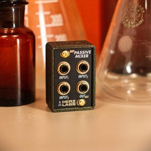 Può includere: Un mixer passivo nero e oro con quattro jack di ingresso e uno di uscita. Il testo "OHP PASSIVE MIXER" è stampato sopra i jack e il logo Herz Labs è in basso. Il mixer è rettangolare e ha una finitura strutturata.
