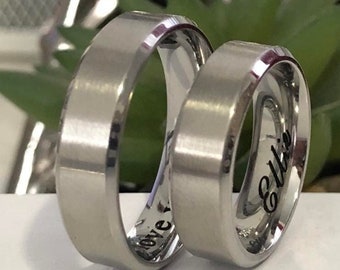 May include: Two silver wedding bands with beveled edges. The band on the left has the word "love" engraved on it. The band on the right has the name "Ellie" engraved on it.