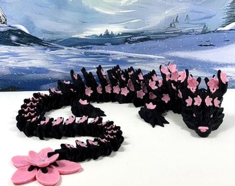 May include: A black and pink origami dragon with a long tail and pink flower accents. The dragon is posed with its head raised and its tail curled.