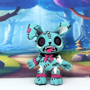 May include: A light blue rabbit figurine with black stitched details, large circular eyes with purple spiral patterns, a wide open mouth with visible teeth and a pink tongue, a black bow tie, and pink wrist and ankle bands on a white base.