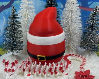 May include: A red and white Santa Claus ornament with a black belt and a white and red garland around it. The ornament is surrounded by miniature white Christmas trees.
