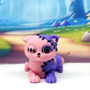 May include: A small cat figurine featuring a split-color design of light pink and purple. The cat has black stitched details across its face and body, one button-style eye, and a small open mouth showing two tiny white fangs on the lower jaw.