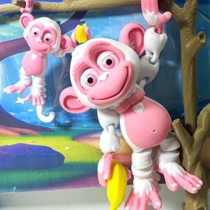 May include: Two white and pink monkey figurines hanging from a brown branch and a tree. The monkeys have pink faces, ears, hands, and feet. One monkey holds a yellow banana. The figurines have a cartoonish design.