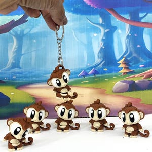 May include: A collection of brown monkey figurines with large white eyes and a keychain. The monkeys have a cartoonish design. One monkey is attached to a silver keychain. The background features a whimsical forest scene.