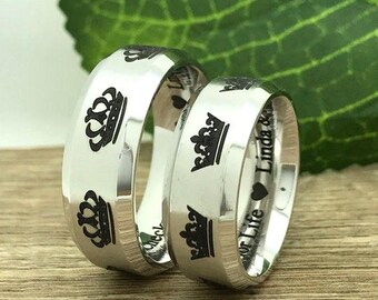May include: Two silver wedding bands with black crown and text engravings. The band on the left has the text "Our Life" and the band on the right has the text "Linda &"