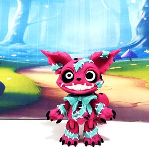 May include: A small, vibrant pink creature figurine with large white eyes, a wide toothy grin, and pointed ears. The figure is decorated with light blue patches and white stitched details across its body, including a bow at the neck and clawed feet.