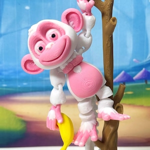 May include: A white and pink articulated monkey toy hangs from a brown branch, holding a yellow banana. The monkey has pink ears, a pink face, and pink hands and feet. The background features a beach scene with blue water and umbrellas.