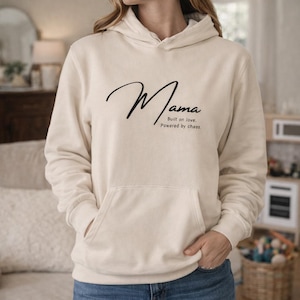 Könnte beinhalten: Cremefarbener Kapuzenpullover mit dem Wort "Mama" in einem stilvollen Schriftzug auf der Brust. Darunter steht der Text "Built on love. Powered by chaos." Der Hoodie hat eine Vordertasche und eine Kapuze.