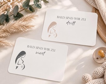May include: Two rectangular white cards with rounded corners featuring a minimalist illustration of a pregnant person and the German text BALD SIND WIR ZU dritt and BALD SIND WIR ZU viert, with a small heart icon on the belly of each figure.