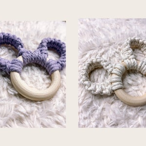 May include: Two wooden teething rings with knotted rope in purple and white. The rings are on a white fuzzy surface.