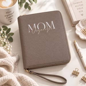Könnte beinhalten: Ein taupefarbener Organizer mit Reißverschluss und Handschlaufe, auf dessen Vorderseite der weiße Schriftzug "MOM Organizer" aufgedruckt ist. Das rechteckige Etui hat eine strukturierte Oberfläche und einen goldfarbenen Reißverschluss zur Aufbewahrung.