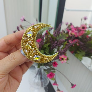 Könnte beinhalten: Eine goldene Halbmond-Brosche mit klaren und gelben Strasssteinen. Die Brosche wird in einer Hand vor einem verschwommenen Hintergrund aus rosa Blumen gehalten.