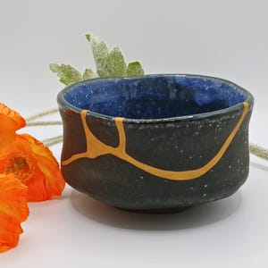 Könnte beinhalten: Eine schwarze Keramikschale mit blauem Innenraum und goldfarbenen Kintsugi-Reparaturlinien. Die Schale hat eine gesprenkelte Textur. Die Schale steht vor einem weißen Hintergrund mit orangefarbenen Blumen und grünem Laub.