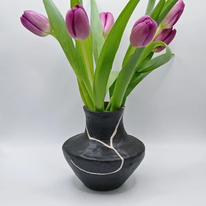 May include: A black ceramic vase with a white, crack-like design, holding a bouquet of purple tulips with green stems and leaves. The vase has a rounded body and a narrow neck, creating a striking contrast with the vibrant flowers.