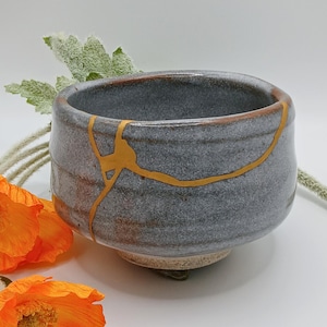 May include: A small, round, gray ceramic bowl featuring a prominent gold-colored repair line running across its exterior surface. The bowl has a textured, speckled finish and a thick rim, showcasing a repair technique that highlights the cracks.