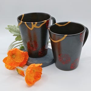May include: Two black ceramic mugs with gold accents and red floral designs. The mugs have a Kintsugi-inspired repair aesthetic. Bright orange poppies and greenery are arranged near the mugs, creating a decorative display.