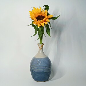 May include: A single bright yellow sunflower with a dark brown center and green leaves sits inside a ceramic vase. The vase has a rounded body with a textured, ribbed surface, featuring a two-tone blue glaze that transitions from light to dark.