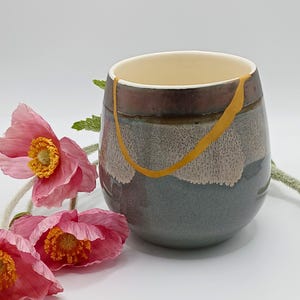 May include: A handmade ceramic cup with a Kintsugi repair. The cup has a gradient of colours, including brown, grey, and blue, with a gold-coloured repair line. Pink flowers with yellow centres are in the foreground.