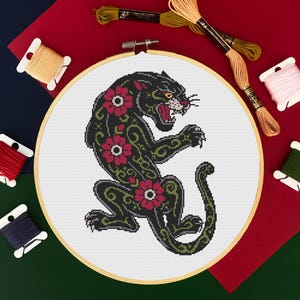 Peut inclure: Un cercle de broderie au point de croix avec un motif de panthère noire orné de fleurs rouges. La panthère est entourée de volutes vertes. L'arrière-plan comprend des bobines de fil de différentes couleurs, dont le rouge, le jaune et le vert.
