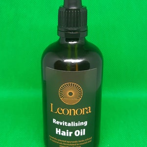 May include: A brown glass bottle with a black dropper top and a white label. The label says "Leonora Revitalising Hair Oil" and includes the text "Carefully selected ayurvedic herbs infused in natural oils formulated to nourish and strengthen the hair. Suitable for all hair types. 100 mls."