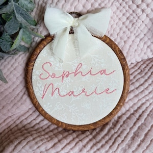 May include: A circular wooden embroidery hoop frame containing cream-colored fabric with a subtle white floral pattern. The name Sophia Marie is embroidered in pink cursive thread across the center, topped with a cream-colored fabric bow.