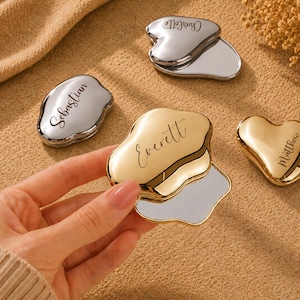 May include: A selection of personalised compact mirrors in silver and gold. The mirrors have various shapes, including a heart and irregular forms. Each mirror is engraved with a name in elegant script. The background is a neutral tan colour.