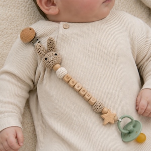 Könnte beinhalten: Ein Schnullerhalter mit einem gehäkelten beigen Hasenkopf, Holzperlen und Buchstabenperlen, die den Namen Mason bilden. Der Halter verfügt über einen runden Holzclip an einem Ende und einen salbeigrünen Schnuller mit gelbem Saugteil.