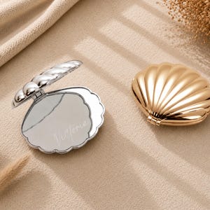Pode incluir: Dois espelhos de bolso em forma de concha, um prateado e outro dourado. O espelho prateado está aberto, revelando uma superfície refletora com o nome Victoria gravado em letra cursiva. Ambos os espelhos apresentam um design festonado.