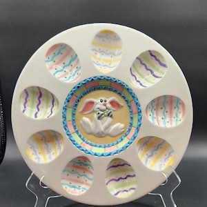 Peut inclure: Plat en céramique blanche pour œufs de Pâques avec 10 indentations en forme d'œuf. Chaque indentation est décorée d'un motif rayé de couleur pastel différent. Un lapin blanc avec un nœud papillon vert se trouve au centre de l'assiette.