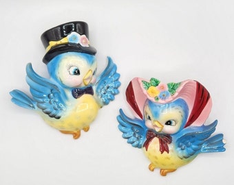 May include: Two blue bird figurines with yellow bellies and painted details. One bird is wearing a black top hat with a pink flower and the other is wearing a pink and red hat with flowers.