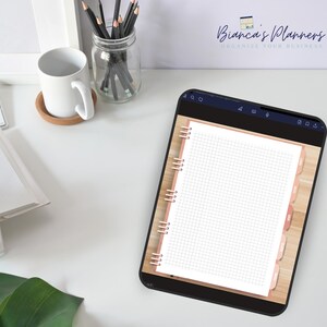 Puede incluir: Una aplicaci&oacute;n de agenda digital en la pantalla de una tableta, que muestra una p&aacute;gina en blanco con un patr&oacute;n de cuadr&iacute;cula. La agenda tiene una encuadernaci&oacute;n en espiral de oro rosa y pesta&ntilde;as. La aplicaci&oacute;n est&aacute; abierta en un escritorio blanco con una taza, l&aacute;pices y una planta.