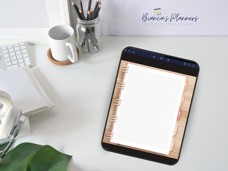Puede incluir: Una aplicaci&oacute;n de agenda digital en la pantalla de una tableta, que muestra una p&aacute;gina en blanco con un patr&oacute;n de cuadr&iacute;cula. La agenda tiene una encuadernaci&oacute;n en espiral de oro rosa y pesta&ntilde;as. La aplicaci&oacute;n est&aacute; abierta en un escritorio blanco con una taza, l&aacute;pices y una planta.
