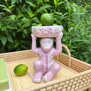 May include: A pink ceramic monkey figurine holding a white bowl with pink floral designs. The bowl is filled with green limes. The monkey wears a vest with a matching floral pattern. The figurine sits on a woven tray.