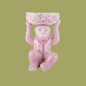 May include: A pink ceramic monkey figurine holding a small bowl above its head. The bowl is white with a pink floral pattern. The monkey is wearing a vest with a white and pink floral design.