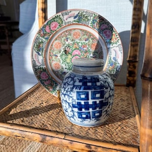 May include: A round ceramic plate featuring a colorful floral and bird pattern with pink, green, and orange accents, positioned behind a small, round white ceramic jar decorated with blue double happiness symbols and vine motifs.