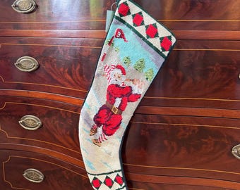 May include: A needlepoint Christmas stocking featuring a scene of Santa Claus playing golf on a light blue course with green trees. The top and toe sections are decorated with a red and white diamond pattern bordered by dark green trim.