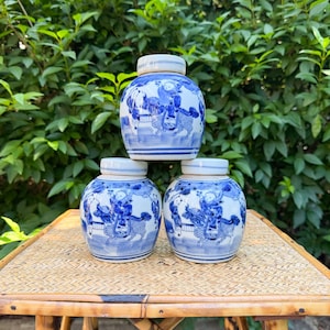 May include: Three white ceramic jars with blue hand-painted illustrations of figures riding mythical creatures. Each jar features a rounded body, a short neck, and a matching lid with horizontal blue bands around the base and rim.