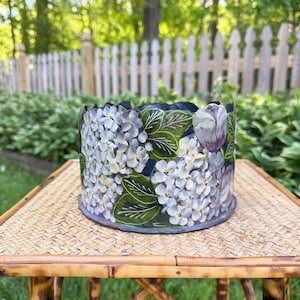 May include: A hand-painted ceramic planter featuring a floral design of white hydrangeas and purple tulips against a dark blue background. The planter sits on a bamboo table, showcasing its decorative appeal.