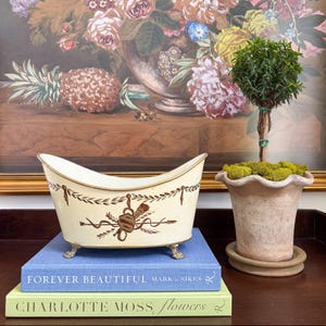 May include: A cream-colored decorative planter with gold trim and bronze-colored accents, resting on a stack of books. The planter has a decorative design and sits on small, ornate feet. A small topiary tree in a terracotta pot is to the right.