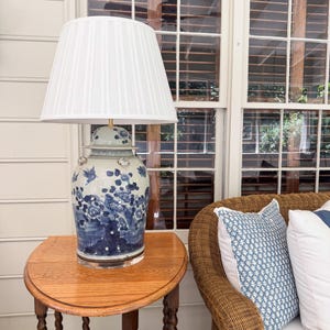 May include: A table lamp featuring a white pleated fabric lampshade and a ceramic ginger jar base. The base is light blue and white, decorated with hand-painted blue floral patterns and bird motifs. It sits on a clear circular acrylic base.