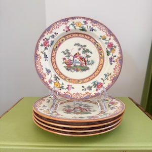 May include: A stack of decorative plates with a floral and bird design. The plates have a cream-colored background with a colorful border of flowers and a central image of birds and a tree. The plates are displayed on a clear stand.