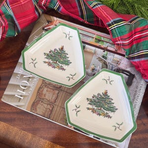 May include: Two triangular ceramic dishes with a cream-colored base and a green rim. Each dish features a central illustration of a decorated Christmas tree surrounded by gifts, accented with small sprigs of mistletoe in the corners of the plates.