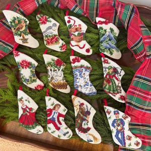 May include: Twelve needlepoint Christmas stockings featuring various holiday designs including birds, swans, a bagpiper, a nutcracker, and figures in period clothing, each with a red hanging loop, arranged on a bed of green pine branches.