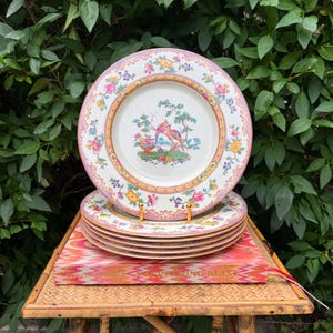 May include: A set of vintage decorative plates with a floral and bird design. The plates have a white background with a pink and gold border. The center of the plates features a colorful bird scene. The plates are stacked on a bamboo table.