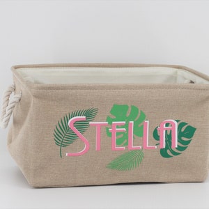 May include: A beige canvas storage bin with rope handles. The bin has a pink and green tropical leaf design and the name "STELLA" printed on the front.