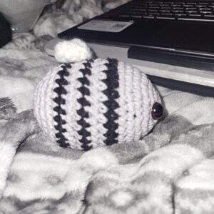 May include: A handmade crochet creature with a white body and black stripes. It has a small white pom-pom on top and brown button eyes. The item is resting on a grey and white patterned blanket.