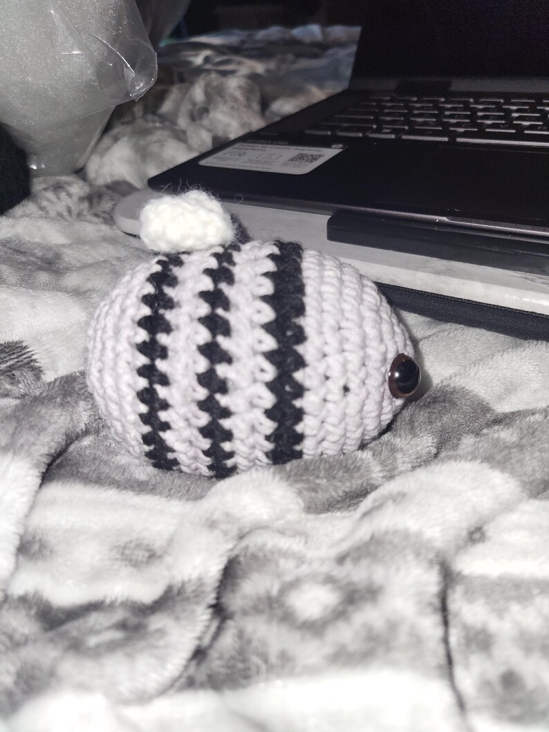 May include: A handmade crochet creature with a white body and black stripes. It has a small white pom-pom on top and brown button eyes. The item is resting on a grey and white patterned blanket.