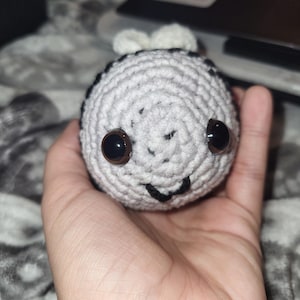 May include: A handmade crochet bee with a grey and white body, black stripes, and large brown eyes. The bee has a smiling face and small white wings. The toy is held in a hand.